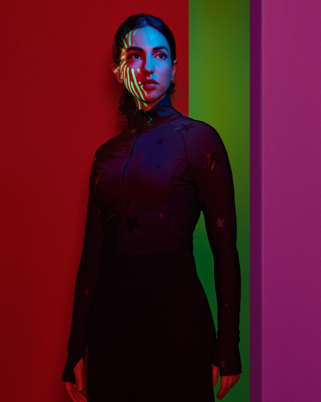 Portrait of a woman in a black turtleneck and skirt stands against a vibrant multicolor wall with a striped light pattern on her face.